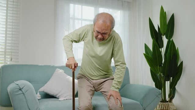 Elderly man suffering from knee pain ache holding handle of cane, senior old man typing to stand up from sofa alone with walking cane stick to walk at home, retirement medical healthcare concept
