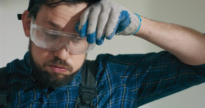Close-up portrait of face of exhausted tired man in work overalls and baseball cap working on home construction renovation. Man wipes sweaty forehead with gloved hand breathing sigh of relief.