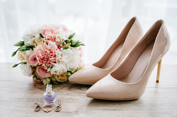 Wedding bouquet of the bride of pink flowers roses and greenery, stylish classic lacquered beige shoes, perfume, earring, and ring lying on wooden background. Bride accessories. Close up. Side view.
