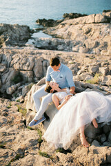 Bride lies on a large stone on the lap of the sitting groom by the sea