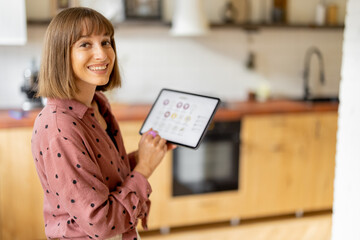 Portrait of a cheerful woman holds a digital tablet with running mobile application for smart home, standing in kitchen of modern apartment. Controling smart devices with a digital tablet - Powered by Adobe