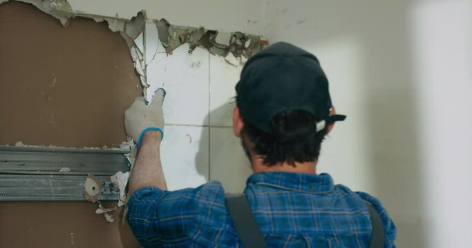 Focused man puts all strength while hammering out tiles in a kitchen bathroom. A man working at home construction site during renovation is doing finishing work with protective gloves.