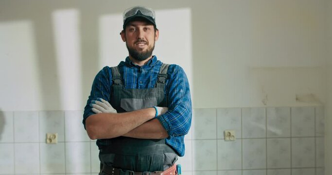 A man working at a construction site dressed in a work overalls baseball cap has arms crossed over chest looks into the camera.