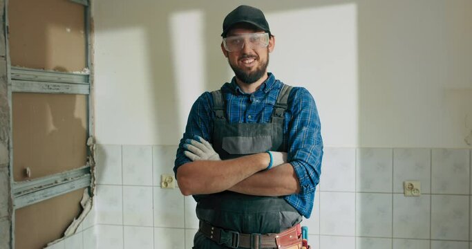 A man working at a construction site dressed in a work overalls baseball cap has arms crossed over chest looks into the camera.