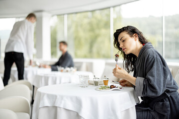 Beautiful woman in bathrobe enjoys delicious meal, sitting in white restaurant of haught cuisine during vacations at luxury spa resort