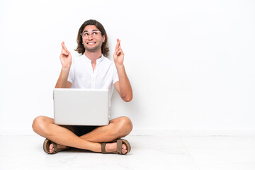 Young handsome man with a laptop sitting on the floor isolated on white background with fingers crossing