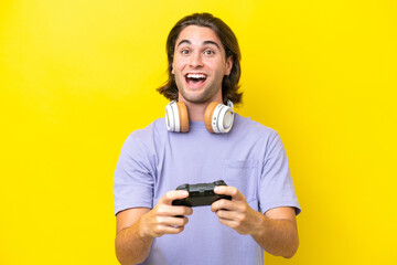 Young handsome caucasian man playing with a video game controller over isolated on yellow background with surprise facial expression