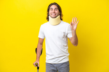Young handsome man wearing neck brace and crutches isolated on yellow background saluting with hand with happy expression