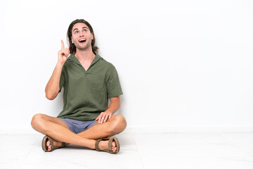 Young handsome man sitting on the floor isolated on white background thinking an idea pointing the finger up