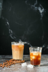 Iced coffee in a tall glass with poured cream, ice cubes and grains on an old rustic wooden table. Cold summer drink with pipes on black background with copy space 