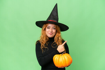 Young caucasian woman costume as witch holding a pumpkin isolated on green screen chroma key background pointing to the side to present a product