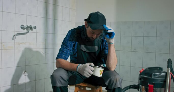 A sad man tired of construction finishing work at home sits with his head bowed leaning his hand against his cheek. The man drinks coffee to wake up gathers strength to continue working.