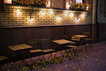 Empty tables and chairs near the cafe on an autumn evening. Light bulbs hang on a brick wall