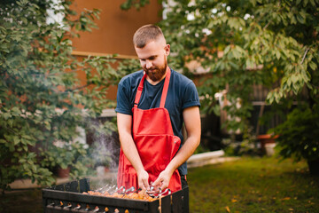 Naklejka na ściany i meble A young man in a blue t-shirt and a red apron with a beard is preparing a barbecue on the grill in the garden near his house. A man fries pork skewers on a metal grill.