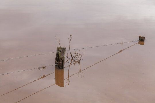 Farm fence covered by muddy brown flood water seen from high ground