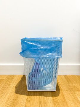 Industrial design garbage can made of white steel with a blue garbage plastic bag inside placed on a wooden floor against white wall background.
