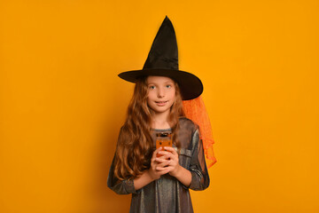 Little witch girl with festive glass standing straight and looking at camera on a yellow background.

Happy surprised girl celebrating Halloween and wearing wizard witch dress costume with black hat.