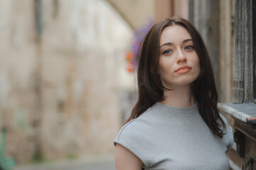 Naklejka premium Beautiful Ukrainian girl with red hair, blue eyes and freckles in a grey summer dress leaning with his back against a wall in a cobbled street with street lamps in the old town