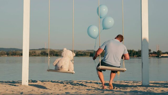 A lonely man together with a teddy bear and inflatable balls sit on a swing near a lake. Sad birthday in loneliness. A lonely man.