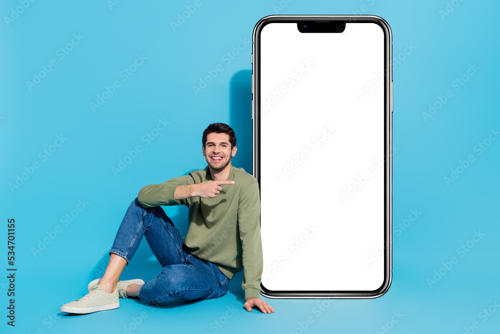 Poster full length photo of cheerful nice man sitting floor indicate finger empty space isolated on blue co
