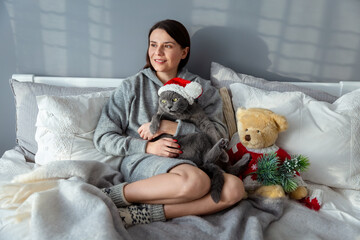 British cat in a  Christmas red hat in the arms of a girl. Girl with a cat and a soft toy on the bed. Bear toy in a Christmas red sweater. New Year theme .Rest in bed
