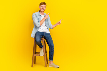 Full size photo of young smiling handsome guy sitting chair finger directing empty space recommend ad isolated on yellow color background