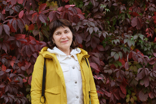 Portrait of an elderly woman against a wall of red autumn vine leaves. Mature woman in a yellow jacket smiles and looks at the camera. City walks, space for text.