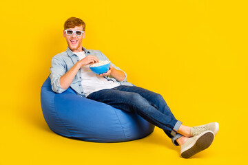 Fototapeta na wymiar Photo of attractive impressed man wear jeans shirt vr eyewear sitting beanbag eating pop-corn empty space isolated yellow color background