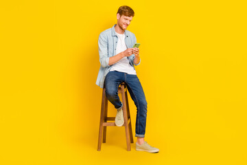 Photo of positive attractive guy dressed denim shirt chatting modern device sitting chair isolated yellow color background
