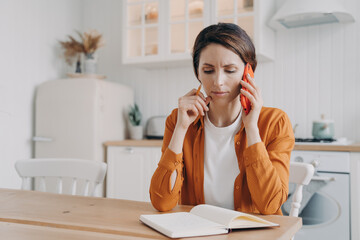 Nervous frowning woman making phone call customer service, sitting at table with notebook in kitchen