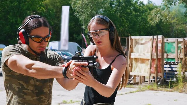 Female caucasian shooting instructor correcting posture of muscular caucasian bearded man. Protective gear - headphones and glasses. Medium outdoor shot. High quality 4k footage