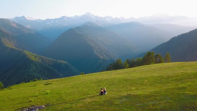 Two young girls or women sit on grass in valley and watch high beautiful mountains. Aerial photography from drone. Georgia Khishkili. Tourists look into distance at beautiful mountain landscape . life