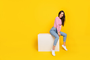 Portrait of cute girlish woman long hairstyle wear striped shirt jeans white shoes sitting on cube isolated on yellow color background