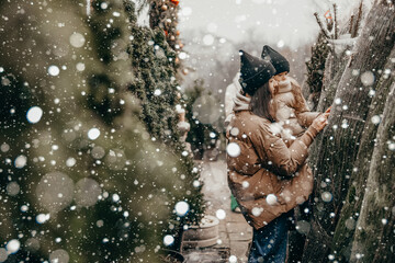 Portrait of a young mother with daughter in her arms, they are smiling and rejoicing in the snowfall. Choosing and buying a Christmas tree at the Christmas market