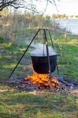 A black metal cauldron on a tripod hangs over a fire, over a cauldron of steam from cooking food, in a forest on a summer day
