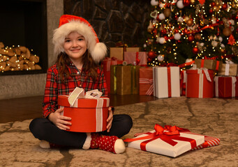 Obraz na płótnie Canvas A happy girl in a santa hat and christmas stockings sits with a gift box on the background of a christmas tree and a fireplace. Christmas mood