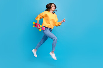 Fototapeta na wymiar Full length photo of pretty charming lady wear yellow sweater jumping high holding longboard empty space isolated blue color background