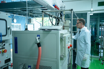 Side shot of Caucasian biochemical scientist with lab coat, gloves and glasses monitoring rotational vaporizer. Expert cannabis researcher checking flask and tube in the process of CBD oil extraction.