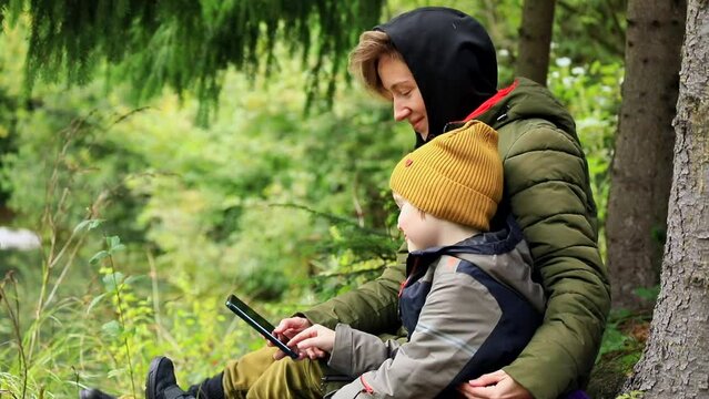 Young smiling mother with happy little boy son using smartphone resting in mountains forest near the lake enjoying beautiful autumn nature. Internet connecting 5g. Traveling family lifestyle concept.