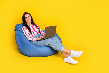 Full size photo of positive cheerful girl striped shirt jeans sit in pouf with laptop chatting...
