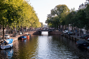 Obraz premium canal con agua y embarcaciones y árboles a los lados y puente al fondo en amsterdam, países bajos, holanda