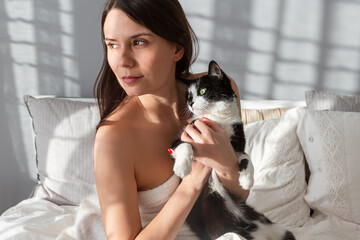 Portrait of a beautiful girl with a cat. Bedroom background. The girl hugs the cat and looks away. Morning in bed.