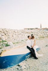 Bride stands next to groom sitting on an upside down fishing boat on the shore