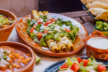 Flautas de maíz con ensalada de lechuga, tomate cebolla y bañadas de crema de vaca, y queso, en un plato de barro tradicional mexicano, en una mesa rustica de madera con crema, tamales y menudo de res