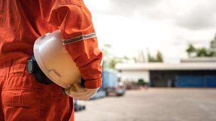 Obraz premium An operation worker in orange coverall is holding white safety helmet or hardhat with factory place as background. Safe working practice in the industrial scene photo, close-up and selective focus.