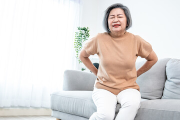 An agonizing senior woman experiencing back pain while sitting on her sofa at home and holding her...
