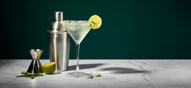 Martini glass with cocktail or mocktail and lime wedge, shaker and jigger on dark background
