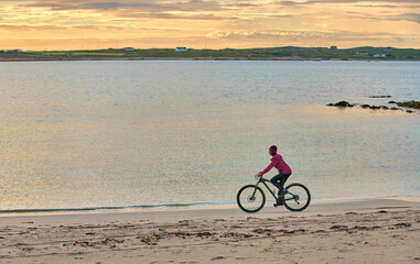 Fototapeta na wymiar nice senior woman on mountain bike, cycling in sunset at the golden sand beach of Sillerna, Grallagh, County Galway, in the western part of the Republic of Ireland