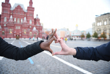 Palms of young people, a European guy and an African girl depicting with their fingers a symbol of love heart on the background of the city outdoors