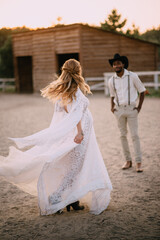 Fototapeta na wymiar Cowboy style wedding. Caucasian bride in boho style dress dances in front of her African groom on ranch.
